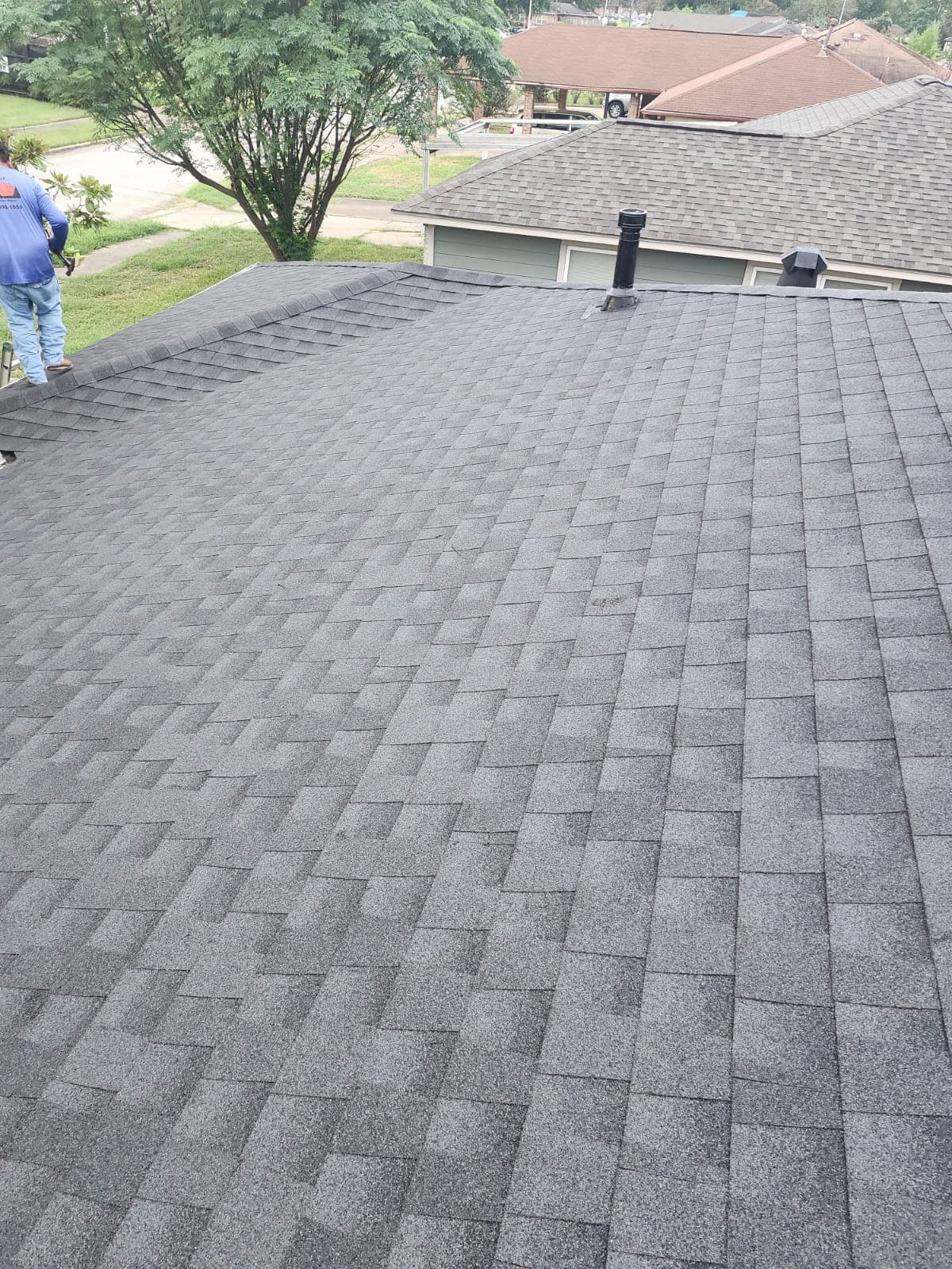 Gray shingled roof with a worker standing on the edge, overlooking a residential neighborhood.